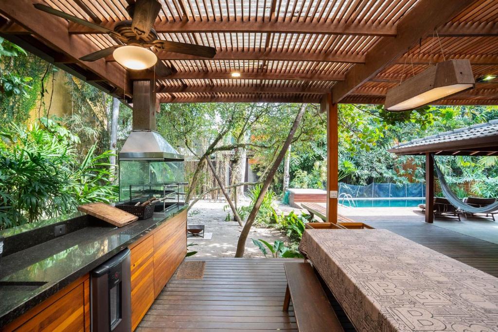 Cozinha externa e área de jantar com pérgola de madeira, ventilador de teto, churrasqueira, mesa comprida e vista para as árvores e a piscina, na propriedade da Casa Tubarão - A Melhor.