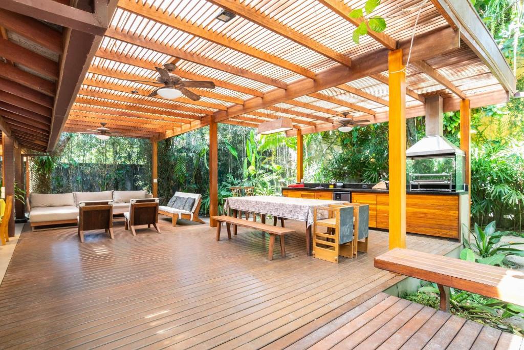 Pátio externo coberto com piso de madeira, mesa de jantar, bancos, área de estar, ventiladores de teto e vegetação ao redor, na propriedade da Casa Tubarão - A Melhor.