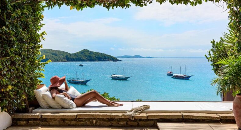 Uma mulher descansa em um sofá-cama sob uma vegetação, observando barcos no oceano com uma ilha ao fundo. O local é o Casas Brancas Boutique Hotel & Spa, um dos resorts em Búzios