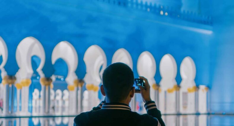 Uma pessoa tira uma foto da grande mesquita de Sheikh Zayed, um construção iluminada em azul, com cúpulas e arcos, à noite. A foto é capa do post de eSIM Abu Dhabi.