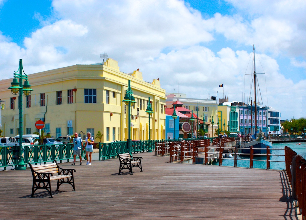 Calçadão de madeira com bancos à beira da água, edifícios coloridos, pessoas caminhando e um veleiro atracado sob um céu azul. Imagem para ilustrar post sobre eSIM Barbados.