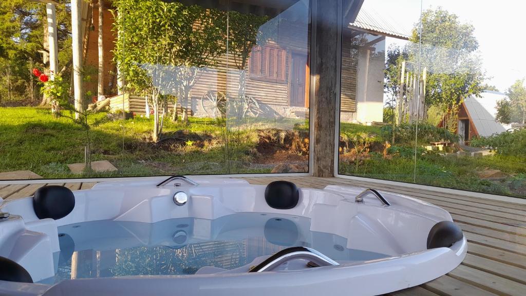 Uma banheira de hidromassagem redonda com água limpa fica em um deck de madeira cercado por paredes de vidro, com vegetação do lado de fora, no Chalé Estância Ouro Verde.