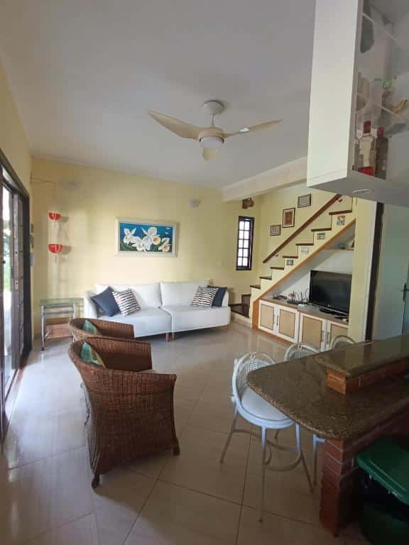 Sala de estar com um sofá branco, cadeiras de vime, TV embaixo da escada e um balcão de café da manhã com duas banquetas, no Chalé Recanto do Barão, para representar aribnb em Boiçucanga.