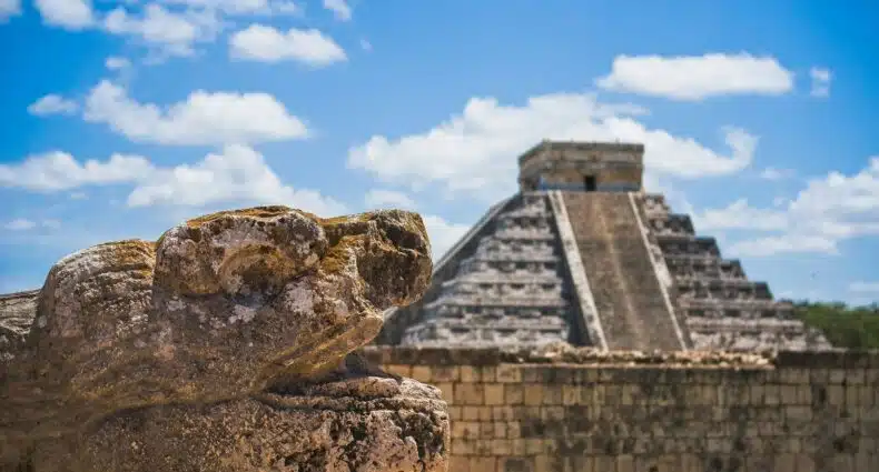 Cabeça de serpente de pedra em primeiro plano com a pirâmide de Chichen Itza ao fundo sob um céu azul com nuvens. Ilustra o eSIM México.
