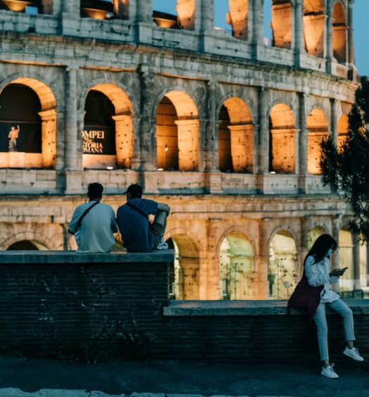 Três pessoas estão sentadas em um muro de tijolos perto do Coliseu iluminado ao anoitecer; uma pessoa está de pé e olha para um telefone. Ilustra o eSIM Europa.