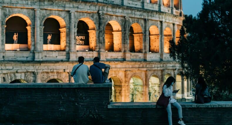 Três pessoas estão sentadas em um muro de tijolos perto do Coliseu iluminado ao anoitecer; uma pessoa está de pé e olha para um telefone. Ilustra o eSIM Europa.