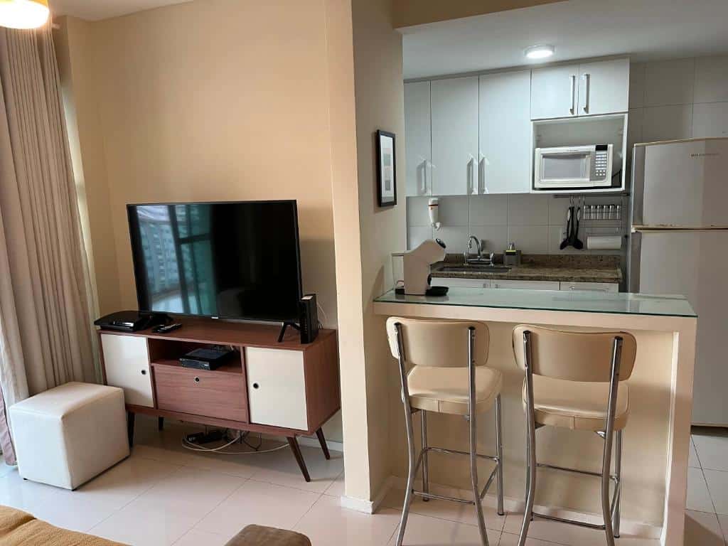 Uma pequena cozinha no Apartamento Bora Bora com armários brancos, uma geladeira e um micro-ondas ao lado de uma sala de estar com uma TV e dois bancos de bar, para representar airbnb na Barra da Tijuca.