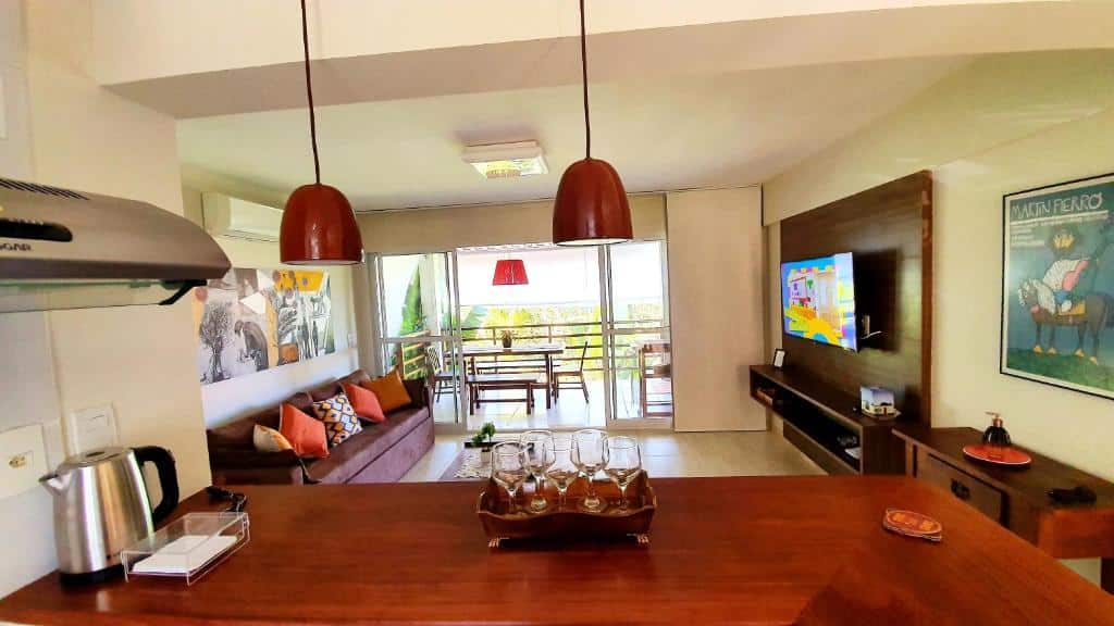 Sala de estar e cozinha no Apartment Delírios abertas com um balcão de madeira, sofás, arte na parede, TV e uma varanda com assentos ao ar livre, para representar airbnb em Imbassaí.