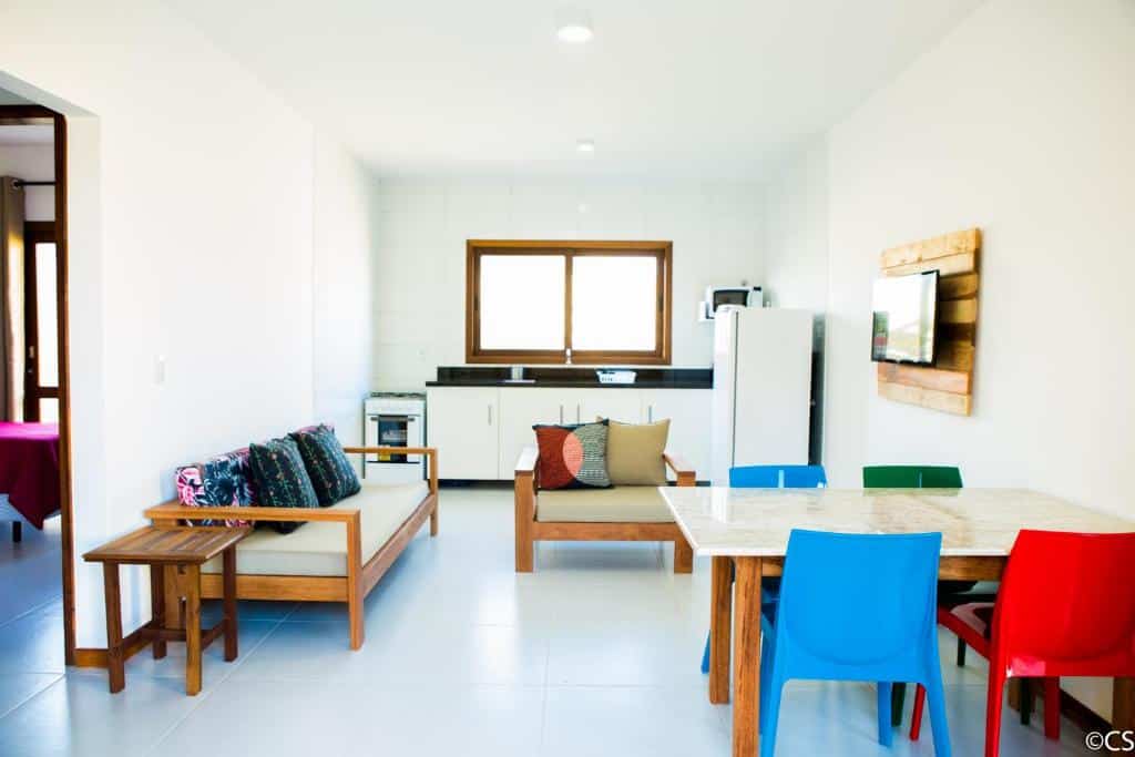 Sala de estar e jantar bem iluminada e minimalista, com cadeiras coloridas, móveis de madeira e uma cozinha ao fundo, na Atlantica Apartamentos Imbassaí, para representar airbnb em Imbassaí.