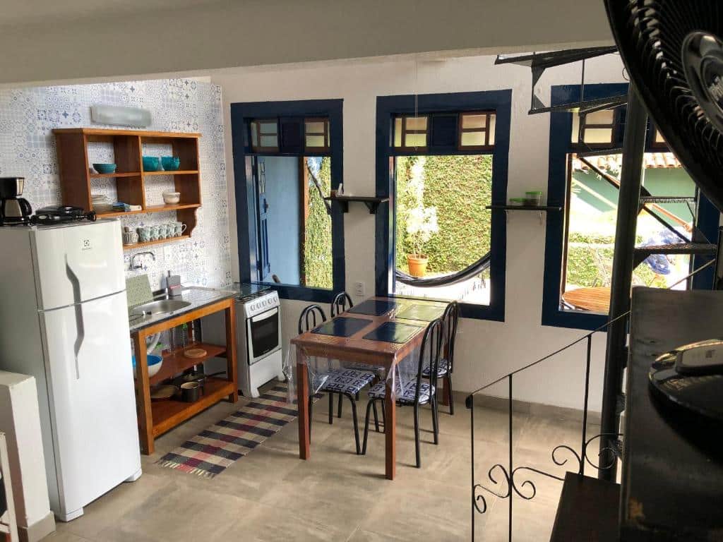 Pequena cozinha e área de jantar com geladeira, fogão, prateleiras de madeira e uma mesa para quatro pessoas ao lado de grandes janelas, no Berrgamota.