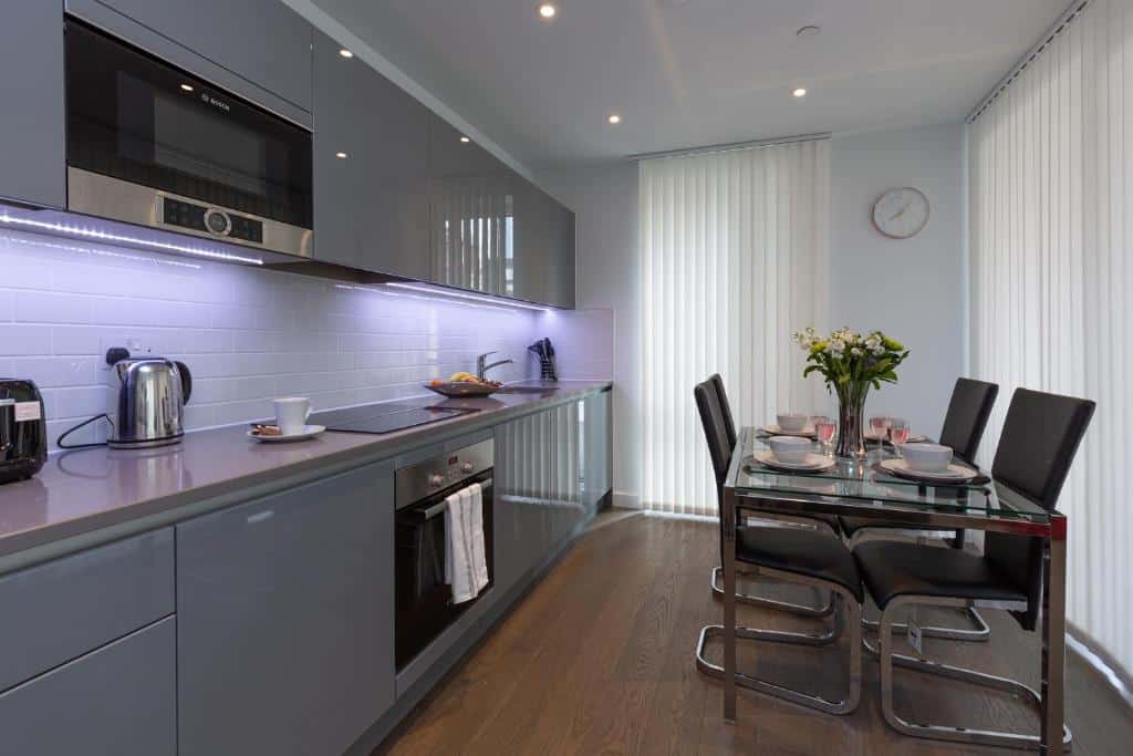Cozinha moderna no Deluxe Central London Apartment com armários cinza, eletrodomésticos embutidos e uma mesa de jantar de vidro para quatro pessoas perto de uma janela com persianas verticais.