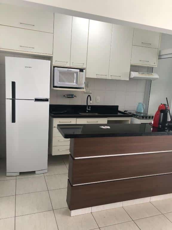 Cozinha moderna com armários brancos, bancada preta, geladeira, micro-ondas, pia, fogão e eletrodomésticos no balcão, no apartamento na Ilha da Madeira Resort com SPA privativo, para representar airbnb na Riviera de São Lourenço.