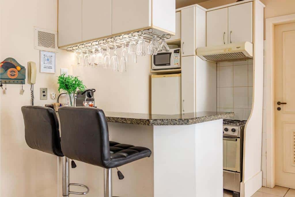 Cozinha pequena com uma barra de café da manhã em granito, duas banquetas pretas, copos pendurados e armários brancos, no apartamento Jurerê Summer - Vista pro Mar.