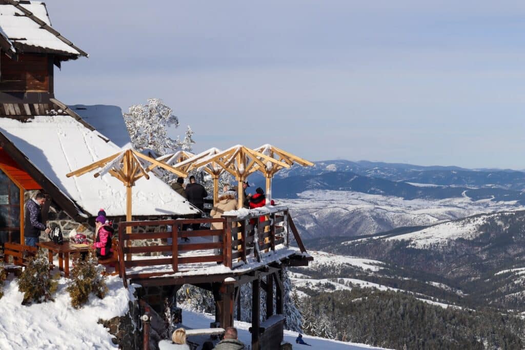 Um deck de madeira com pessoas em roupas de inverno tem vista para uma paisagem de montanhas nevadas sob um céu claro. Ilustra o eSIM Sérvia.