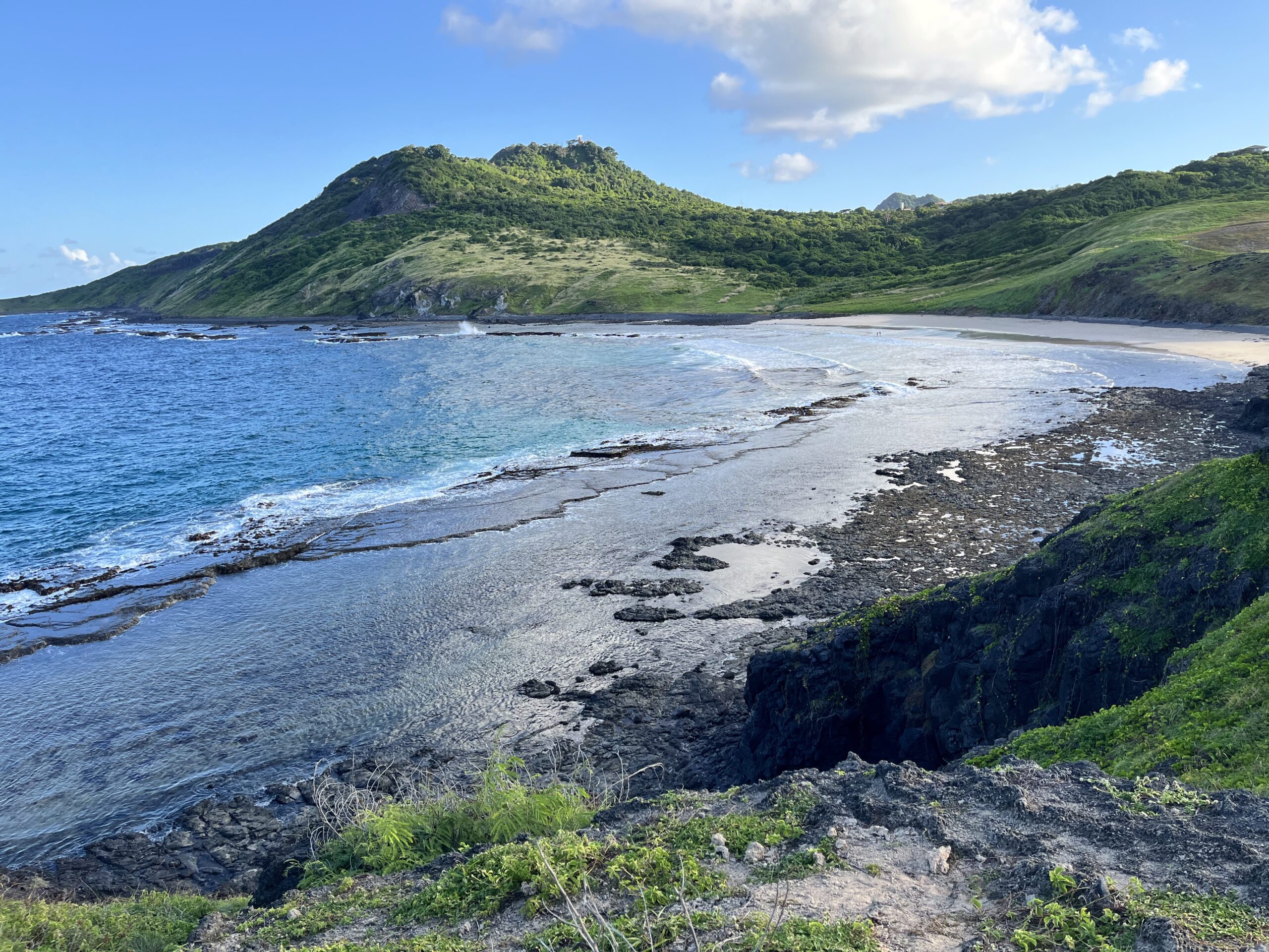 Um litoral rochoso com águas azuis e cristalinas, praia de areia e colinas verdes sob um céu parcialmente nublado.