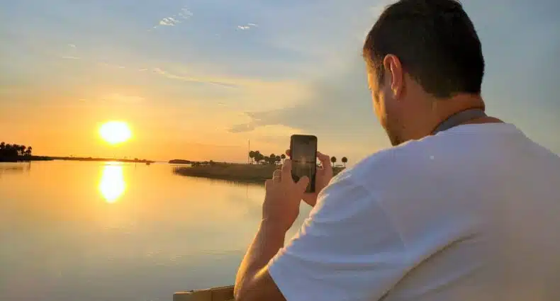 Um homem de camisa branca tira uma foto do pôr do sol sobre águas calmas com seu telefone. Imagem para ilustrar post sobre eSIM Flórida.