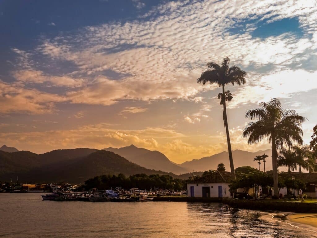 Palmeiras e um pequeno prédio à beira d'água com barcos ancorados, montanhas e um céu nublado ao fundo no pôr do sol, em Paraty.