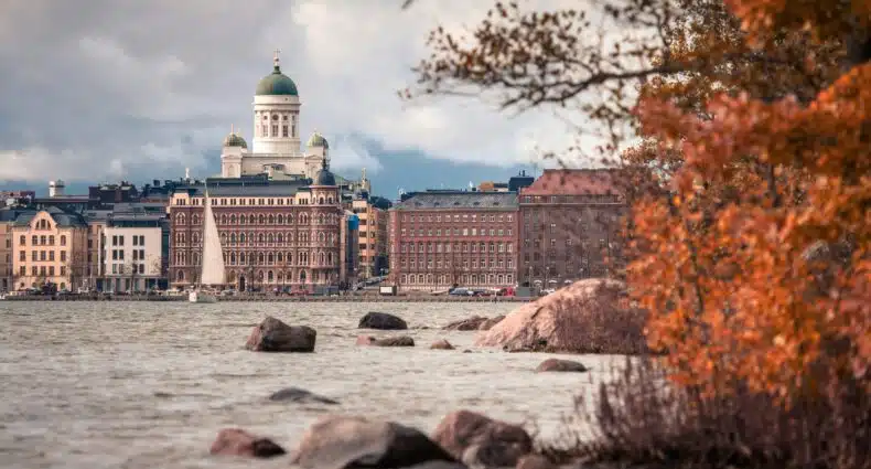 Catedral de Helsinque e edifícios à beira-mar vistos do outro lado da água, com árvores de outono e pedras em primeiro plano.