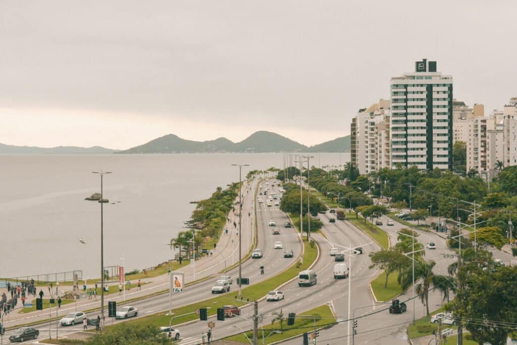 Uma estrada costeira com carros e edifícios de um lado, água e colinas ao fundo sob um céu nublado, em Florianópolis.