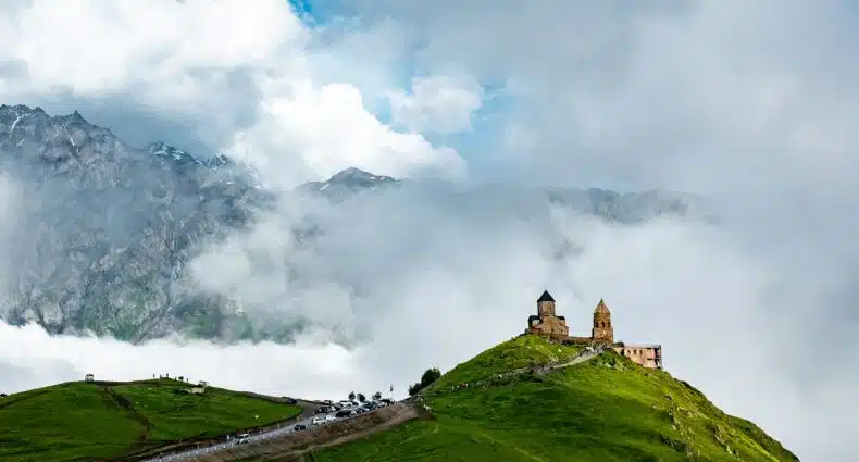 Uma igreja de pedra fica no topo de uma colina verde com montanhas enevoadas e nuvens ao fundo. Ilustra o eSIM Geórgia
