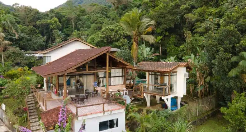 Uma casa com telhado de telhas vermelhas, janelas grandes e um deck, cercada por densas árvores verdes e plantas tropicais, no Haleiwa Chalés e Suítes, para representar pousadas em Prumirim.