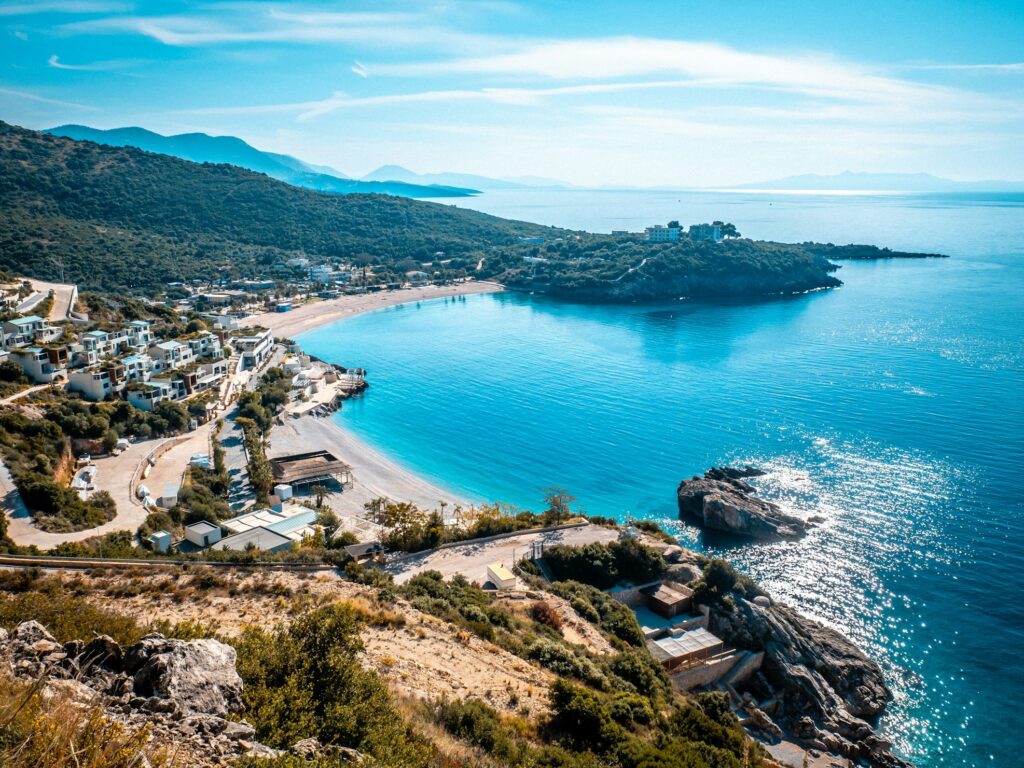 Uma baía costeira com águas azuis e cristalinas, praia de areia e colinas verdes, com edifícios ao longo da costa sob um céu ensolarado. A praia é a Jale Beach, na Albânia. A foto está no post de eSIM Albânia.