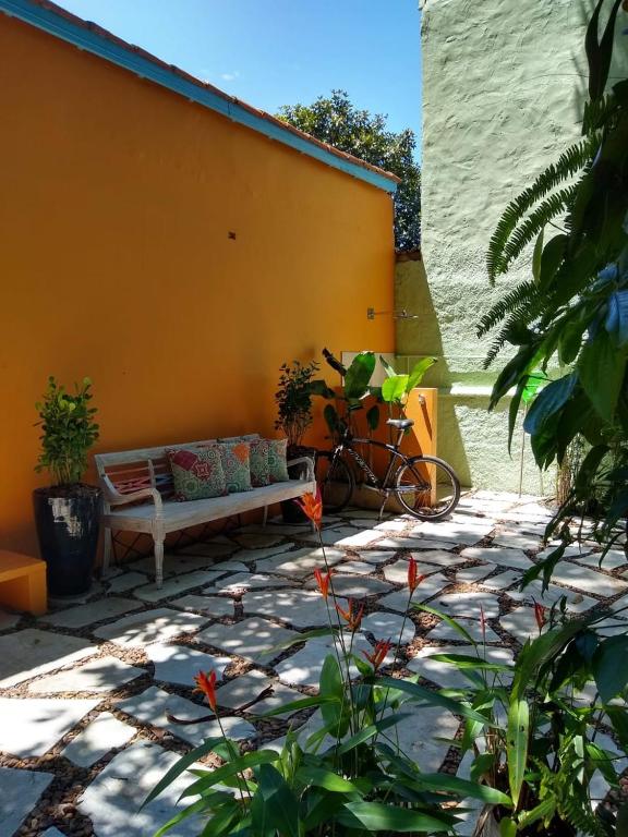 Pátio externo com um banco branco, vasos de plantas, uma bicicleta e ladrilhos de pedra contra paredes amarelas e verdes, nas Suítes Km18praia.