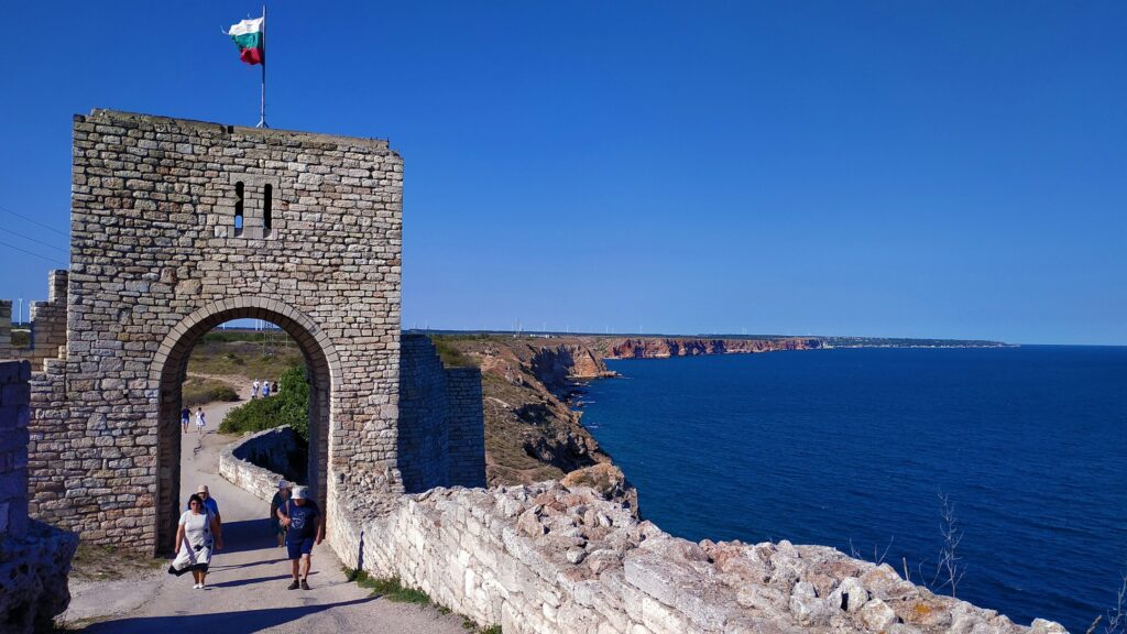 Kaliakra, na Bulgária. Um arco de pedra com a bandeira búlgara tem vista para um penhasco costeiro e para o mar azul, com pessoas caminhando em um caminho abaixo do arco. A foto faz parte do post de eSIM Bulgária.
