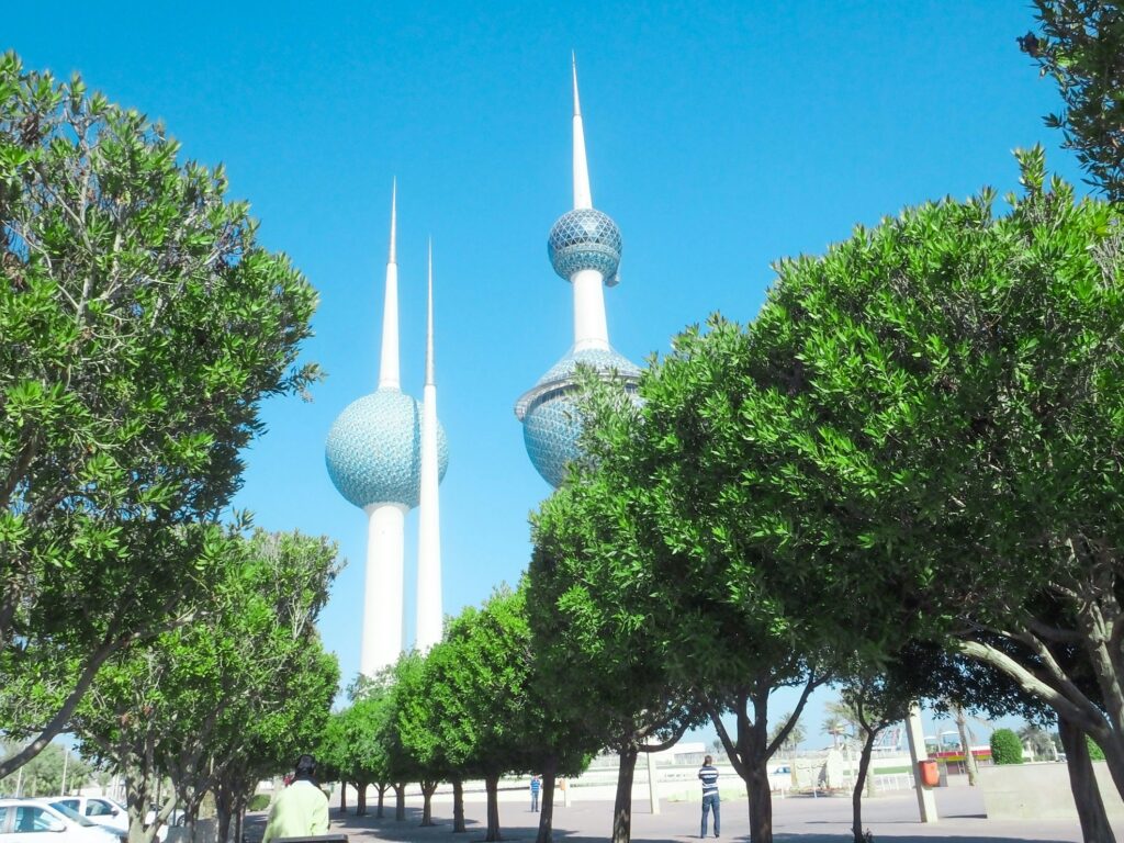 Três Torres Kuwait erguem-se atrás de árvores verdes sob um céu azul claro, com algumas pessoas visíveis perto das árvores. A foto faz parte do post de eSIM Kuwait.