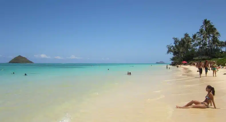 Pessoas relaxam em Lanikai Beach com águas azul-turquesa, palmeiras e um céu azul claro ao fundo. Imagem para ilustrar post sobre eSIM Havaí.