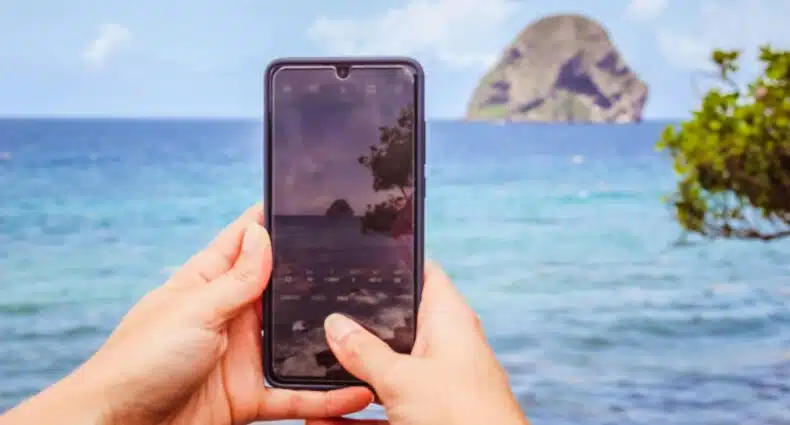Uma pessoa segura um smartphone para fotografar uma ilha ou formação rochosa em um oceano azul sob um céu parcialmente nublado. A foto foi tirada em Le Diamant, na Martinica. A foto é capa do post de eSIM Martinica.