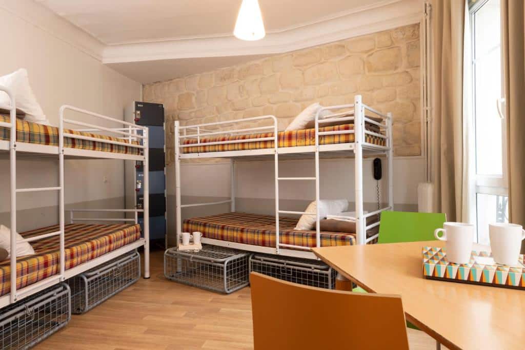 Um quarto com dois beliches de metal, uma mesa com quatro cadeiras, canecas e uma janela que permite a entrada de luz natural, no Le Regent Montmartre by Hiphophostels.