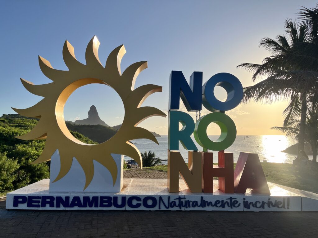Grande placa colorida de "Noronha" com um desenho de sol, palmeiras e o oceano ao fundo ao pôr do sol.