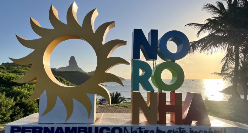 Grande placa colorida de "Noronha" com um desenho de sol, palmeiras e o oceano ao fundo ao pôr do sol.