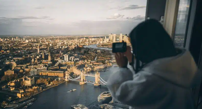 Pessoa com uma jaqueta branca tirando uma foto do horizonte de Londres, incluindo a Tower Bridge, através de uma janela ao pôr do sol. Ilustra o eSIM Inglaterra.