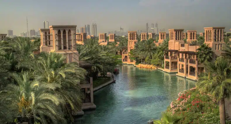 Um canal com águas azul-turquesa corre entre palmeiras e edifícios bege, com o horizonte da cidade visível ao fundo no Madinat Jumeirah. Imagem para ilustrar post sobre eSIM Dubai.