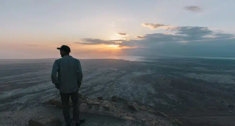 Uma pessoa de boné está em um mirante rochoso, no Masada National Park, em Israel, observando uma vasta paisagem desértica sob um céu nublado de pôr do sol. A foto faz parte do post de eSIM Israel.