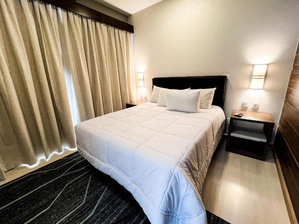 Uma cama bem arrumada com roupa de cama branca em um quarto moderno, ladeada por duas mesinhas de cabeceira e luminárias de parede, no Master Cosmopolitan Moinhos de Vento, para representar onde ficar em Porto Alegre.