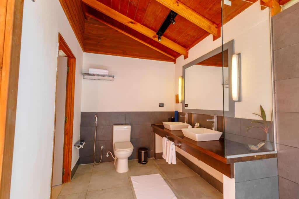 Banheiro moderno no Meeru Island Resort & Spa com vaso sanitário, pias duplas, espelho grande, teto de madeira e paredes e pisos de azulejos cinza.
