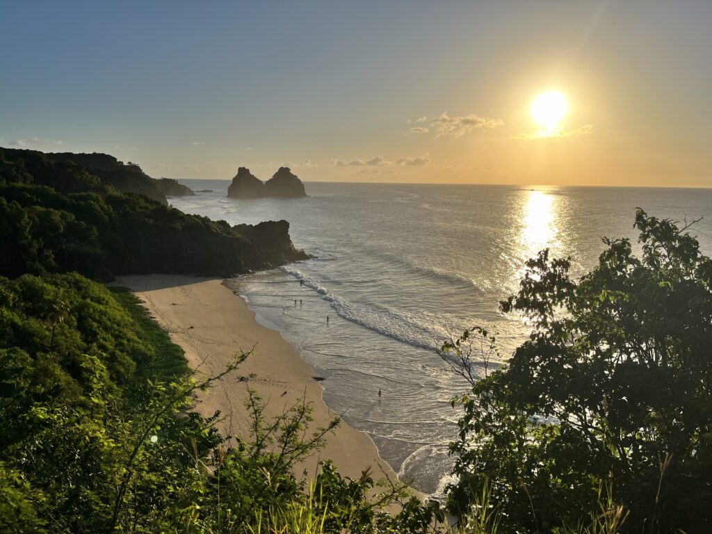 Uma praia de areia com ondas suaves, um litoral rochoso e o sol se pondo sobre o oceano, parcialmente cercada por vegetação.