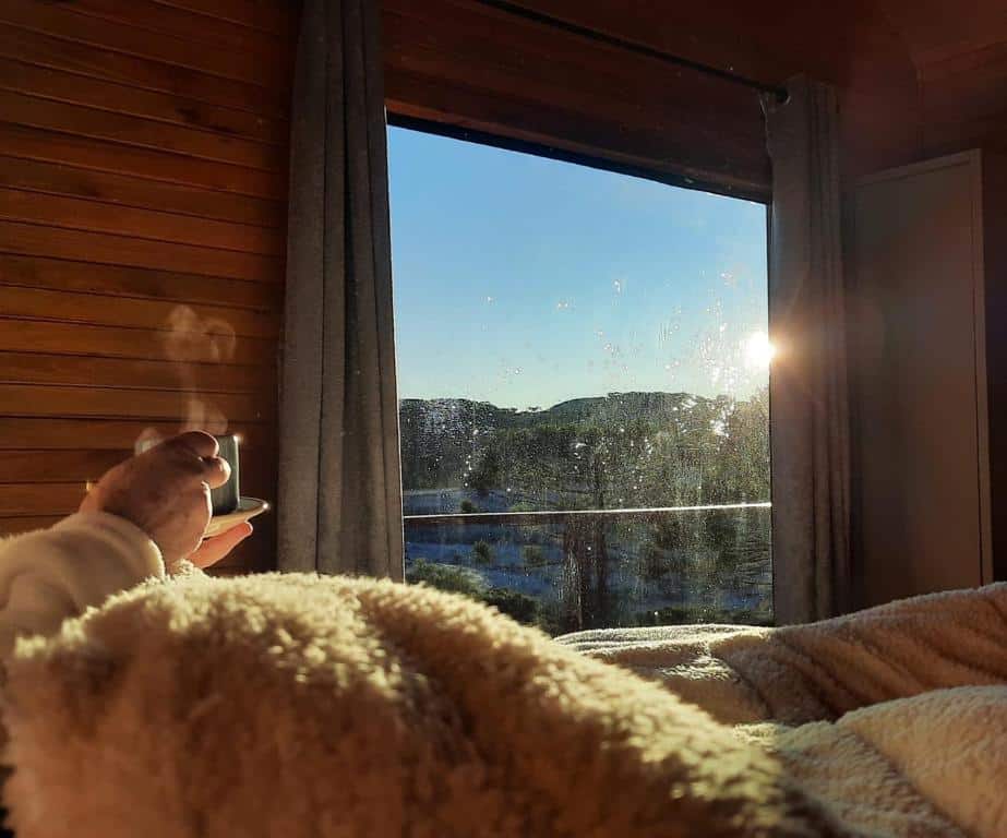 Uma mão segurando uma caneca fumegante perto de uma janela iluminada pelo sol com cortinas, olhando para as colinas e um céu azul claro. O local é a Monte Alegre Pousada, em São Joaquim