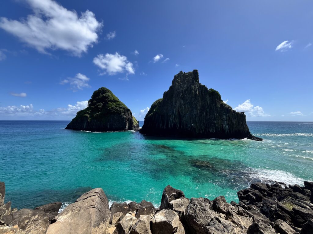 Duas ilhas rochosas cobertas de vegetação erguem-se de águas azul-turquesa cristalinas sob um céu quase ensolarado, vistas de uma costa rochosa.