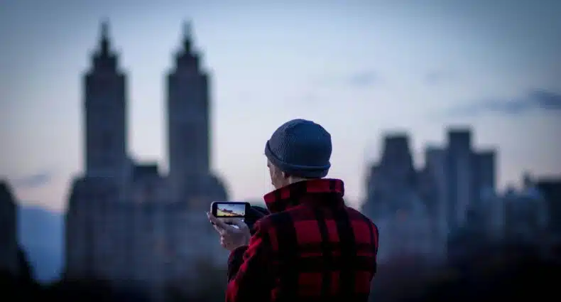 Uma pessoa com uma jaqueta xadrez vermelha e chapéu cinza tira uma foto de prédios em Nova York com um smartphone ao anoitecer. A foto faz parte do post de eSIM América do Norte.