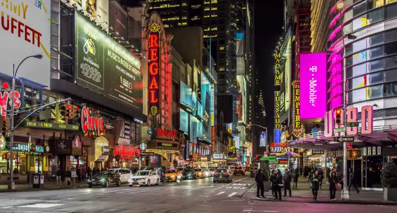 Times Square à noite com letreiros de neon brilhantes, outdoors, lojas, carros e pessoas atravessando a faixa de pedestres. Imagem para ilustrar post sobre eSIM Estados Unidos.