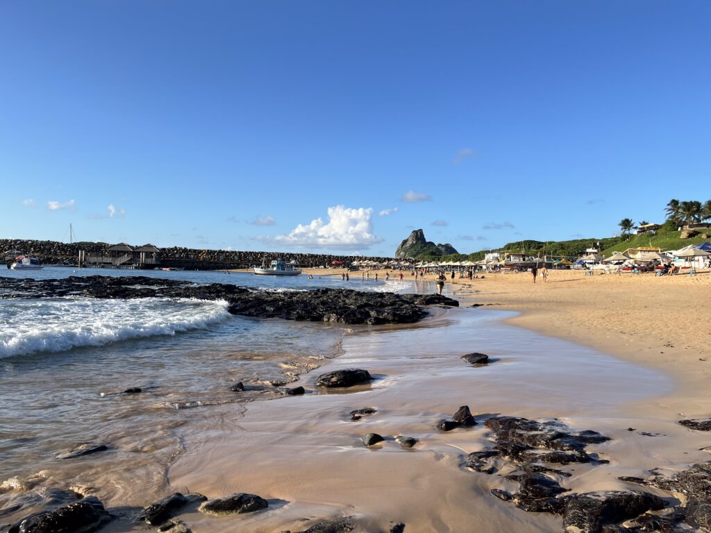 Uma praia de areia com pedras escuras, ondas suaves, pessoas e prédios, com uma colina verde e céu azul ao fundo.