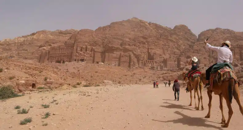Pessoas montadas em camelos tiram uma selfie perto de antigas estruturas escavadas na rocha em penhascos em uma paisagem desértica em Petra, na Jordânia.
