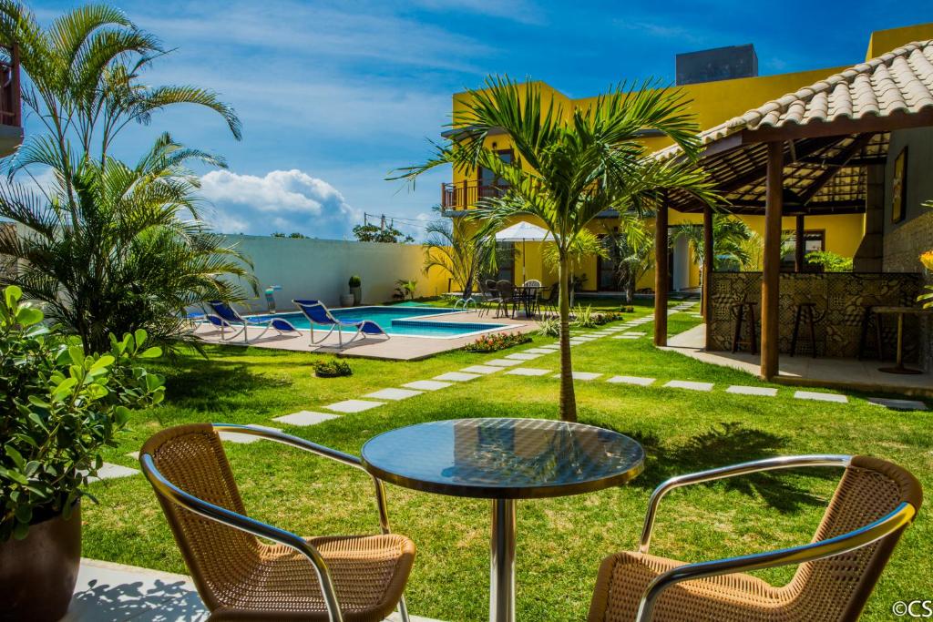 Um quintal ensolarado com piscina, espreguiçadeiras, palmeiras, uma área de pátio e uma pequena mesa com duas cadeiras em primeiro plano, na propriedade do Atlantica Apartamentos Imbassaí.