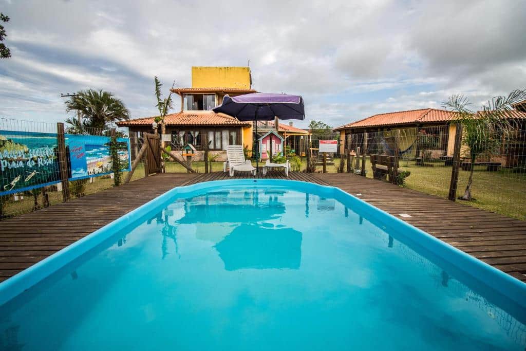 Uma pequena piscina externa com um deck de madeira, espreguiçadeiras e casas com telhados vermelhos ao fundo, sob um céu nublado, na propriedade das Cabans do Rosa.
