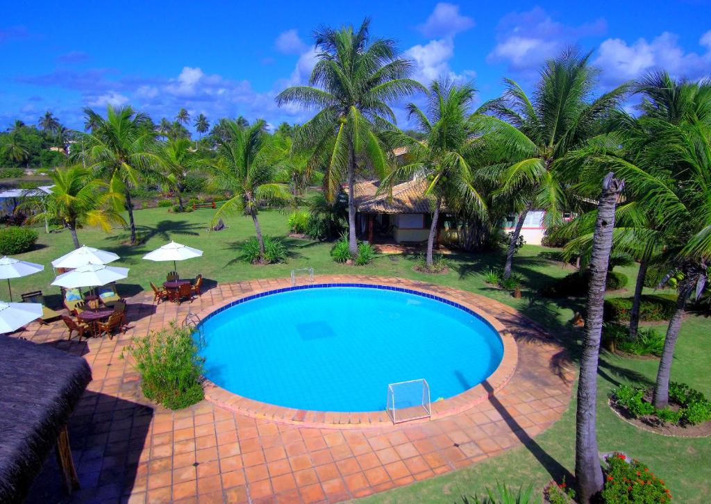 Uma piscina redonda ao ar livre é cercada por palmeiras, grama e espreguiçadeiras com guarda-sóis em um dia ensolarado, na propriedade da Grimms Pousada de Contos na Costa do Sauípe.