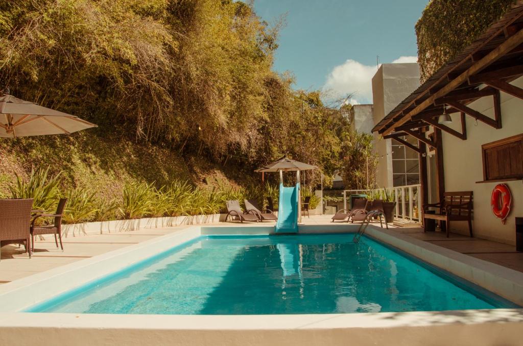 Uma piscina externa retangular com um escorregador azul, espreguiçadeiras, guarda-sóis e vegetação ao fundo, na propriedade do Hotel Marambaia Cabeçudas - frente mar.